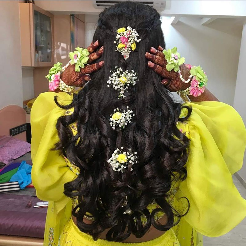 Bridal hair in glossy curls with white floral pins, henna on the hands, and a yellow lehenga sleeve.